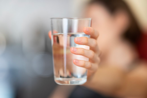 A woman, out of focus, holds a plain glass full of water, in focus, toward the camera.