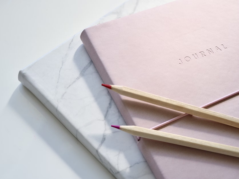 Two journals, one marble-patterened and one light pink with the word "Journal" embossed on it, sit stacked together. Two colored pencils sit on top of them.
