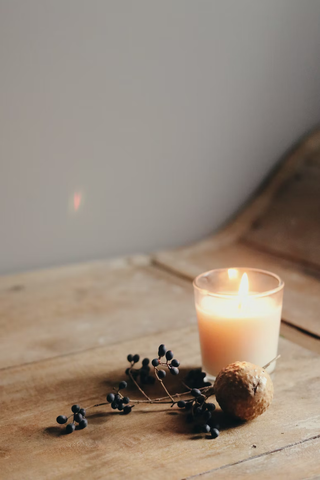 A lit white tumbler candle alongside some berries and a nut on a wooden table.