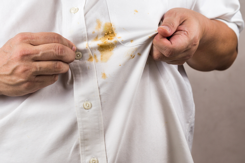 Closeup of a man's white button-up shirt. He holds out a yellow stain near the chest pocket.