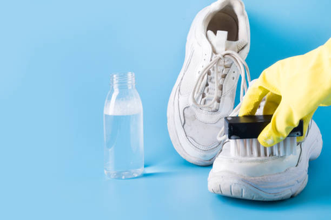 A hand in a yellow latex glove uses a sponge to clean a pair of white sneakers.