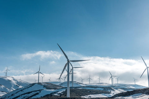 A wide shot of many energy windmills on a snowy hill range. The sky is blue and lightly cloudy.