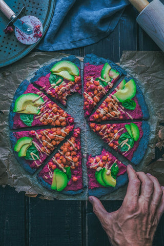 An overhead shot of a purple tart dish topped with various fruits and vegetables, cut into eiht slices. A hand reaches for one half-eaten slice.