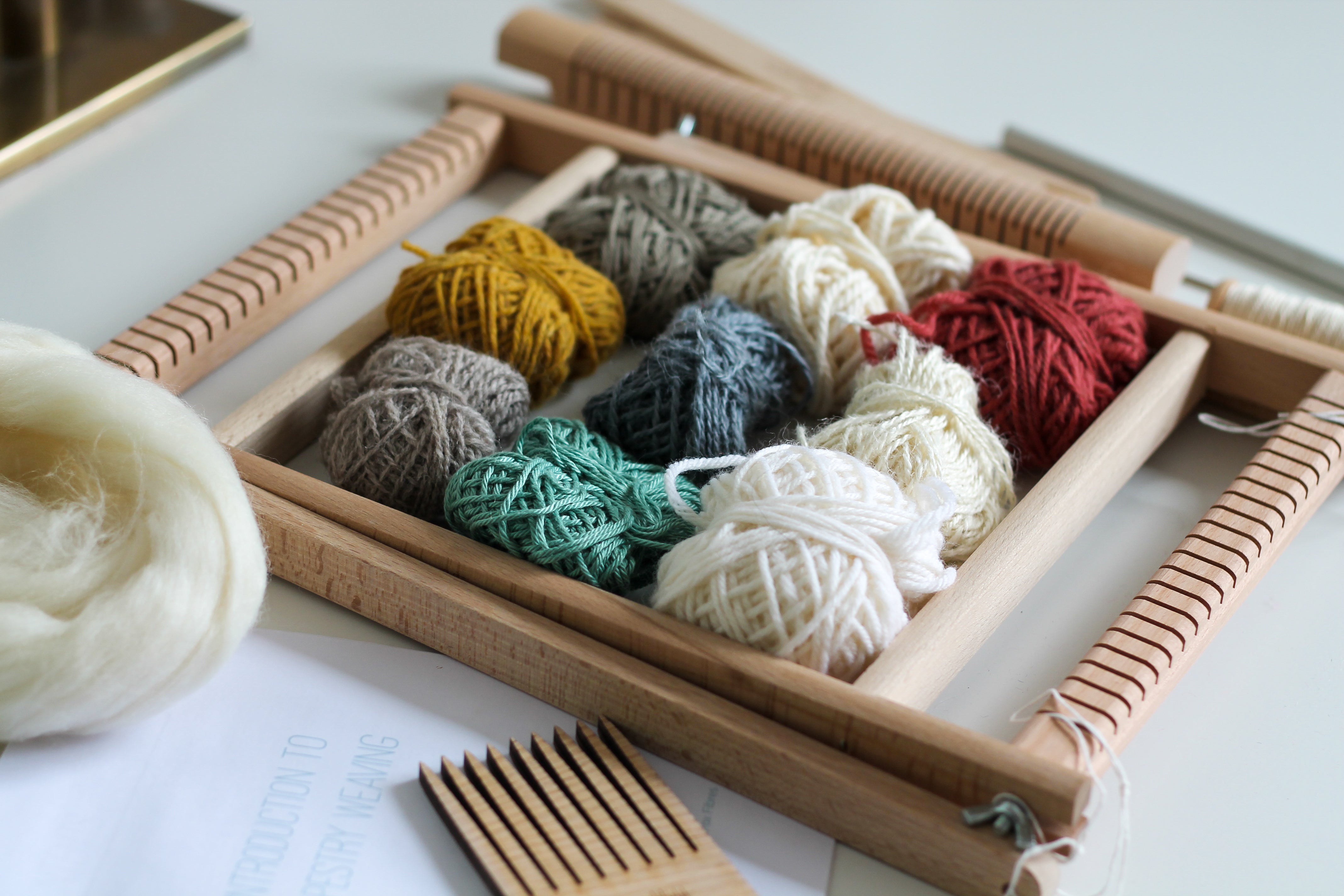 weaving loom, assorted yarn, tools and a booklet are laid out on a white table