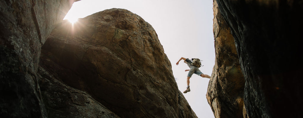 A hiker jumping across large boulders captured from below looking up with the sun beaming through the mountains showing what KAIBAE customers are often found doing in the wild
