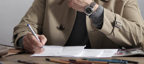 stressed person in beige jacket sitting at desk, writing on piece of paper with pen