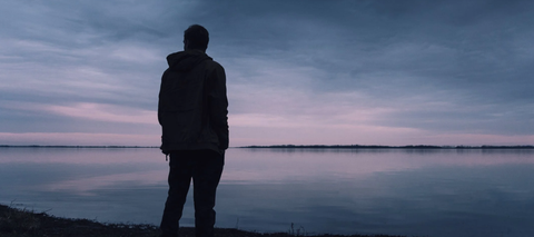 silhouette of person in coat standing at lake edge at dusk and staring at horizon