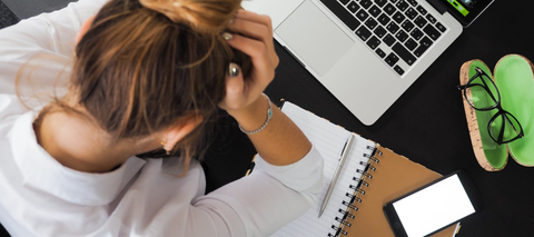 overwhelmed woman holding head in hands in front of laptop and notebook