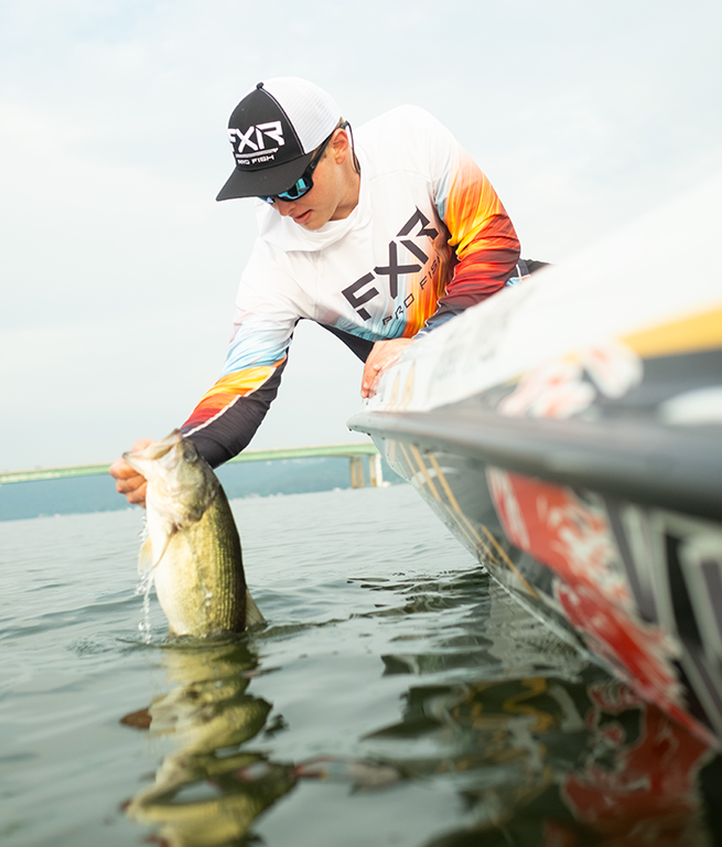 An angler on a bass boat releasing a largemouth bass wearing FXR’s rust bone Tournament Hybrid UPF pullover hoodie.