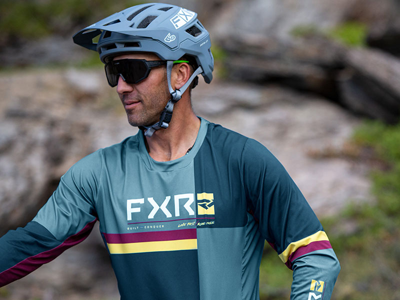 An image of a guy wearing the FXR's Proflex UPF short sleeve jersey in Steel/Sundial pushing his mountain bike