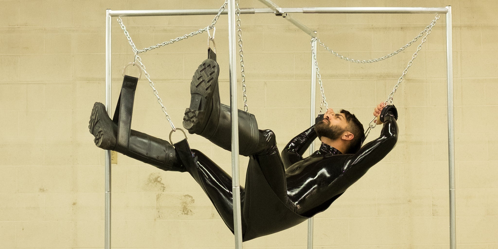 A brunette man with facial hair wearing a black latex catsuit is shown laying in a black leather sex swing with stirrups, held up by chains on a metal frame.