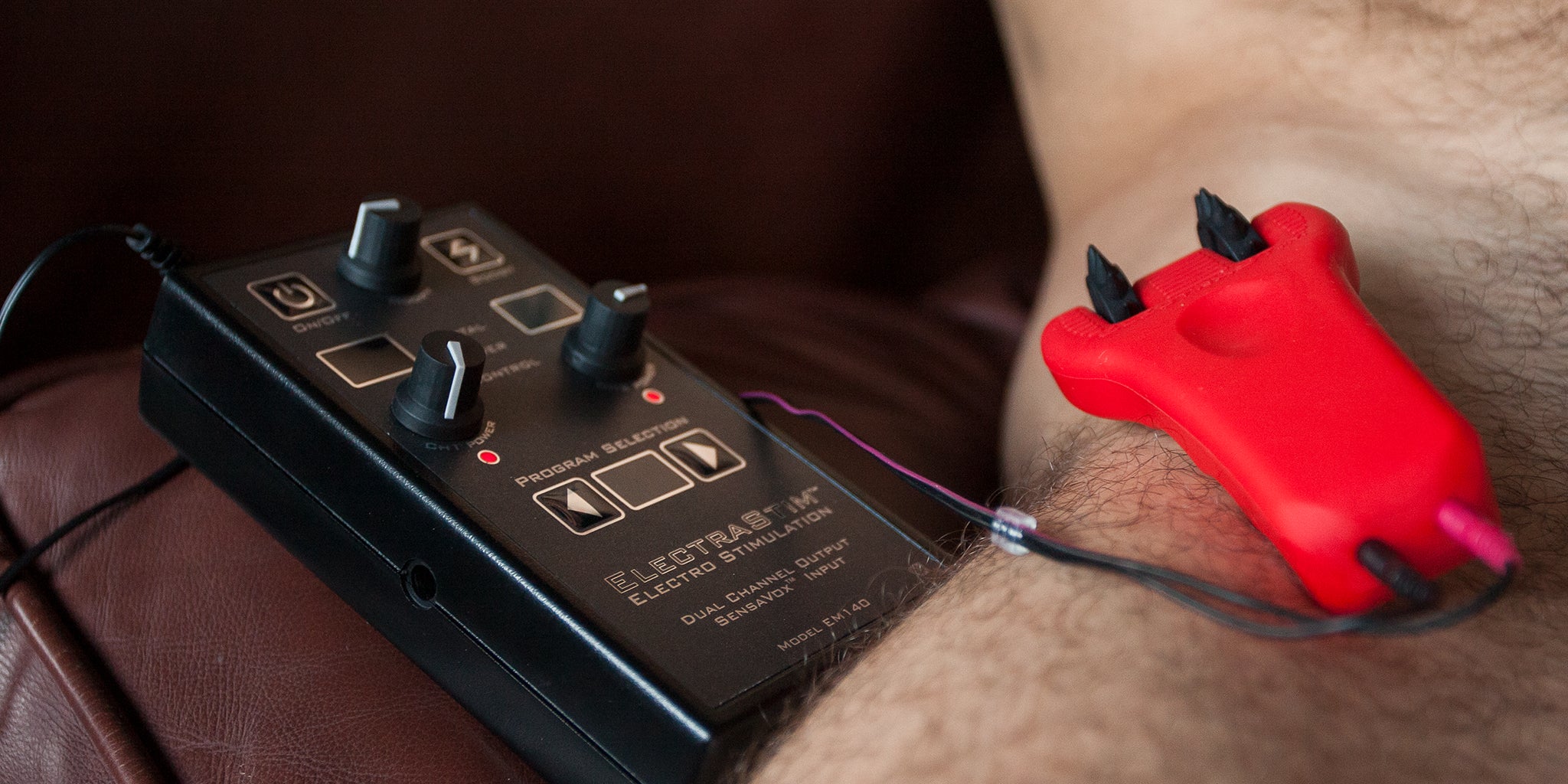 A close-up shows the black Electrastim Sensavox controller plugged into the red and black Electrastim Silicone Fusion Infinity Pinwheel, which rests on a man’s pelvis.