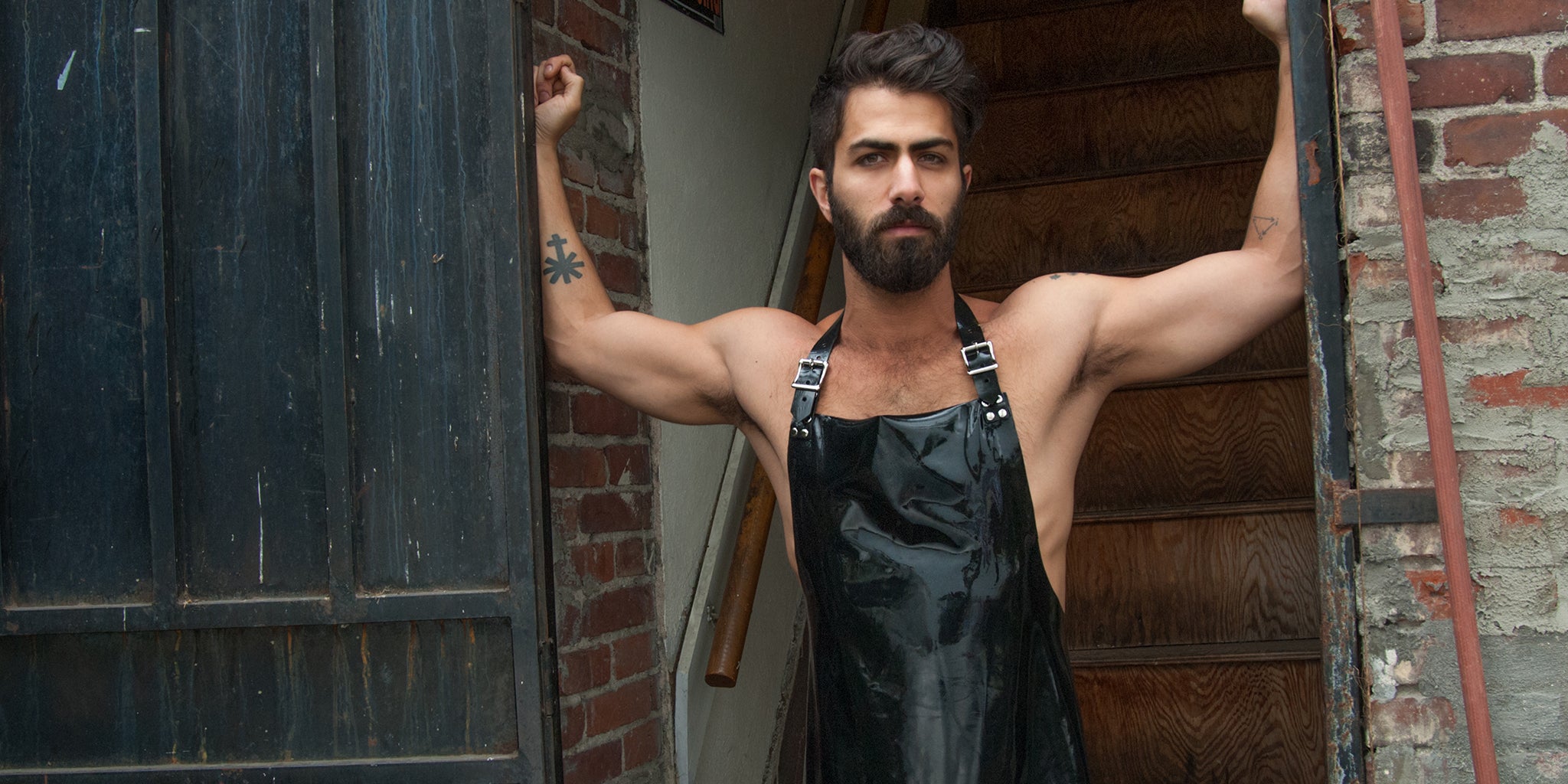 A brunette man is shown from the hips upward, standing in a doorway. He is shirtless and wears the black Rubber Apron from the Stockroom.