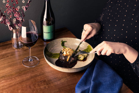 Woman cuts into a steak on a plate, with a bottle of red wine to her right and a glass of wine in front of her