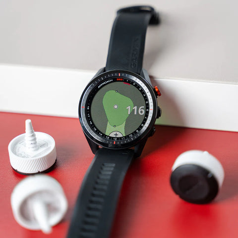 The black Garmin Approach S70 golf watch leaning against a wall on a red floor with CT10 tracking sensors around it