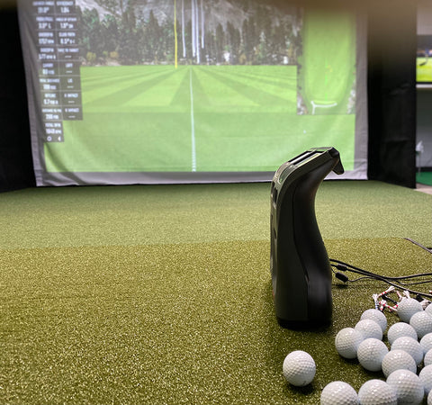 Side view of the Launch Pro and GC3 launch monitors on a golf mat next to some golf balls with the bottom of a simulator impact screen in the background