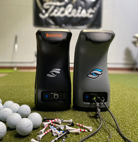 The Bushnell Launch Pr o and Foresight GC3 side by side from the rear in a golf simulator next to golf balls and tees