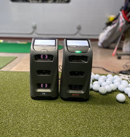 The Bushnell Launch Pro and Foresight Sports GC3 side by side on a golf hitting mat near some golf balls in an indoor golf simulator