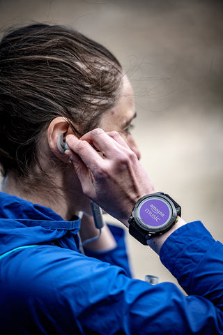 A woman stopping during a run adjusting earbuds with Garmin Enduro 2 GPS smartwatch on her wrist with the Amazon music app showing
