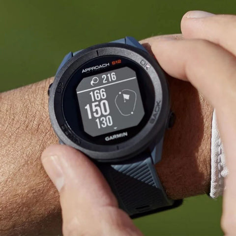A black Garmin Approach S12 golf watch on a wrist with fingers on the buttons showing golf distances on the display