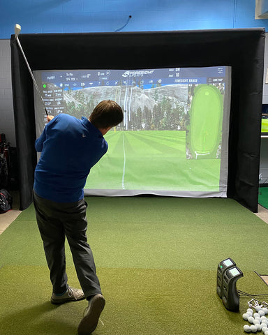 Marc swinging in an indoor golf simulator in front of the impact screen next to the Bushnell Launch Pro and GC3 launch monitors