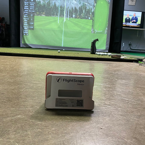 The FlightScope Mevo+ set up in an indoor golf simulator with a golf impact screen with virtual golf software on it and a GC3 launch monitor in the foreground