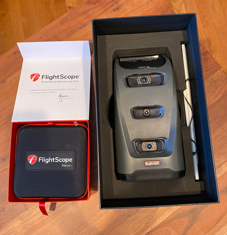 The FlightScope Mevo+ and Bushnell Launch Pro next to each other in their open boxes on a wooden floor