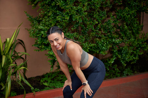 A woman pausing during a run outside with her hands on her knees smiling and wearing a Garmin running watch