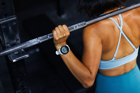 A woman shown from behind getting ready to press a barbell overhead wearing a white Coros Pace 3 sport GPS watch