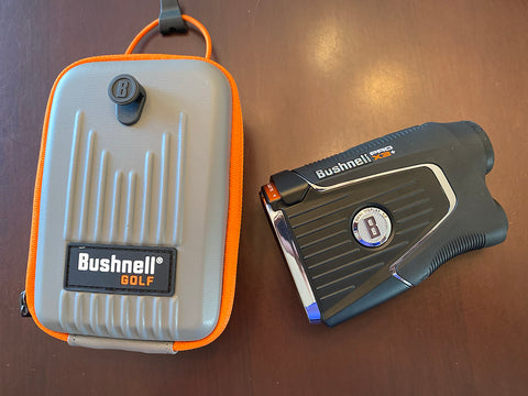 The Bushnell Pro X3+ rangefinder laying on a table next to its gray and orange Bushnell carrying case