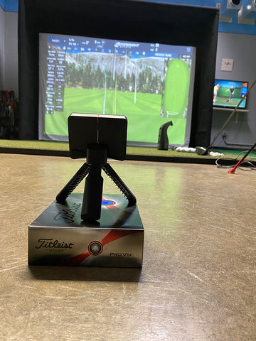 The Garmin Approach R10 sitting on a Titleist golf ball box in an indoor golf simulator studio