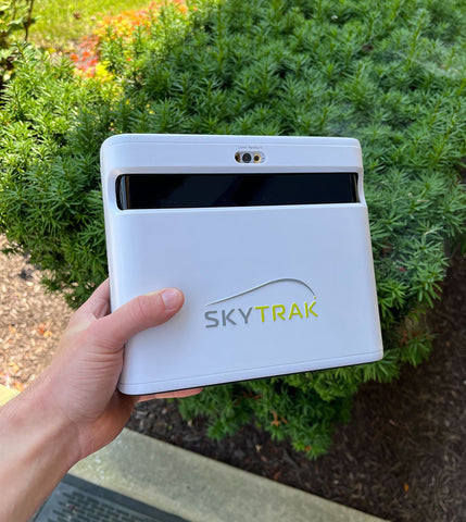 The SkyTrak+ being held in a hand outside over some hedges