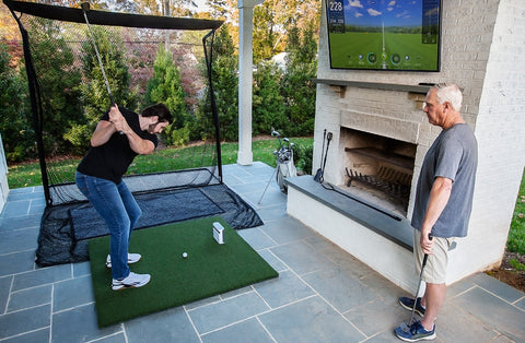 Two golfers, one watching, one swinging on an outdoor golf setup with SkyTrak+, golf hitting net, golf turf, and golf hitting net, with simulation on a TV hanging above a fireplace on the patio