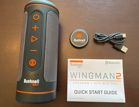 The contents of the Wingman 2 box, including golf GPS speaker, charging cable, remote, and quick start guide laid on a table