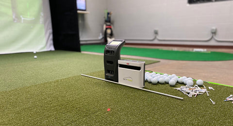 The Launch Pro and SkyTrak Plus side by side on a golf mat next to golf balls and golf tees in an indoor golf simulator