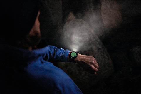 Over-the-shoulder view of a hiker in a rocky area at night looking at a Garmin fenix 7X Pro watch on their wrist with the flashlight illuminated and maps on the display