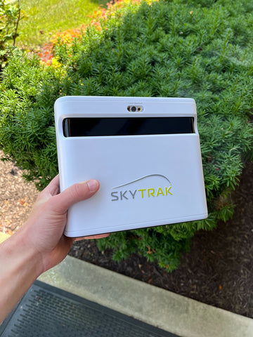The SkyTrak+ launch monitor unit being held in a hand over grass and bushes