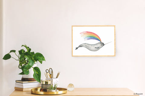A framed print of a narwhal that blows a rainbow out of it's spout. It is in a thin wooden frame hanging on a light pink wall, behind a desk with a plant and brass and glass office supplies.