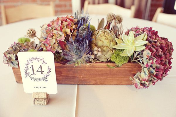 A trough of flowers and a table number placed in the centre of a dining table at a natural wedding