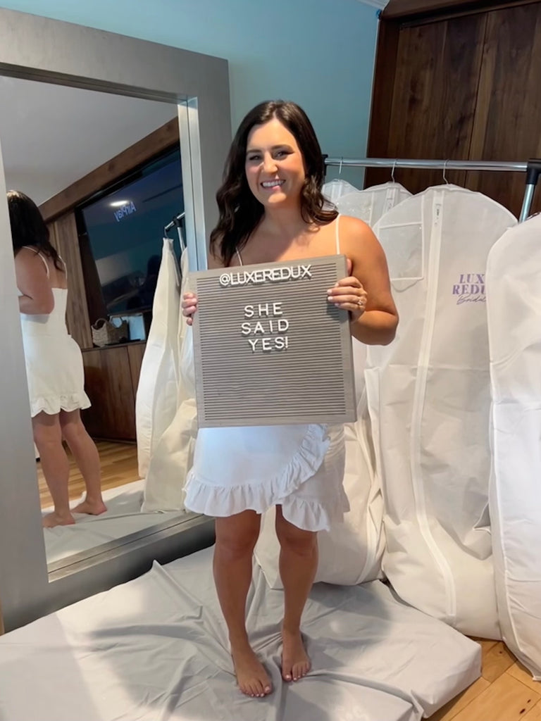 Bride standing in front of wedding garment bags with a sign that says "She said yes!"