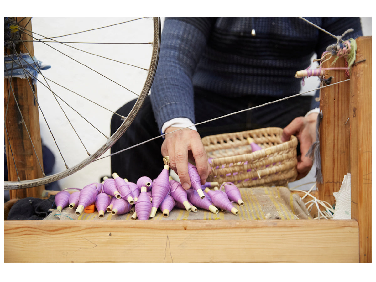 Image of an artisan sitting at a weaving loom and holding a basket of violet colored yarn spools.