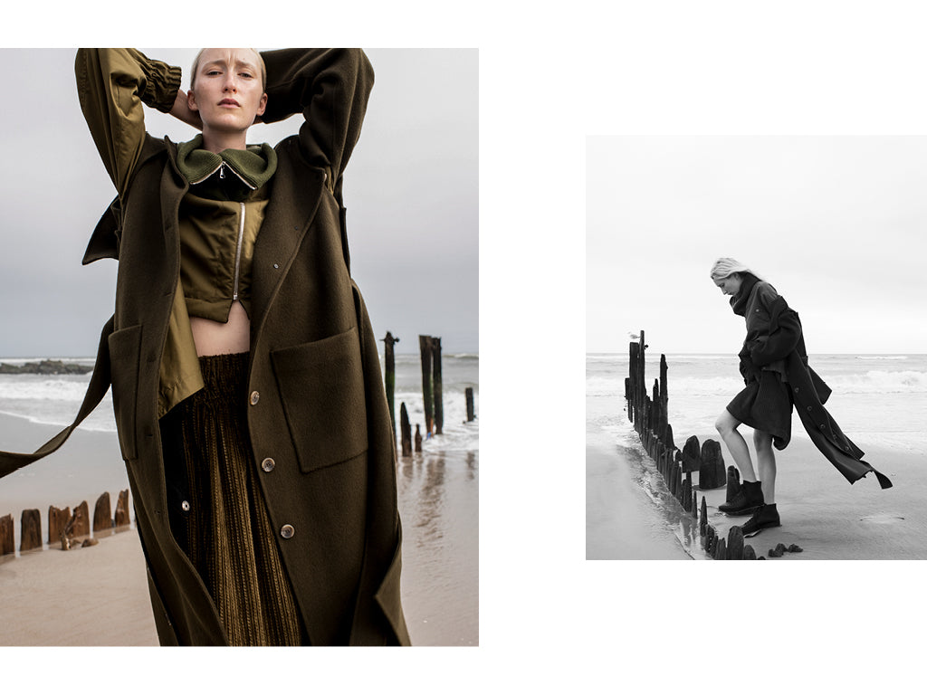 A cropped front view of a woman standing at the beach wearing a long corduroy skirt and zip front jacket by Nehera with a dark Green wool coat by Rejina Pyo. The second image is a Black and White side view of the same Blonde haired woman standing on the oceanfront wearing Black Leather hi-top sneakers by FEIT.