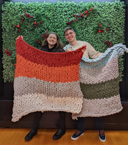 Two people hold up chunky knit blankets in front of a moss wall