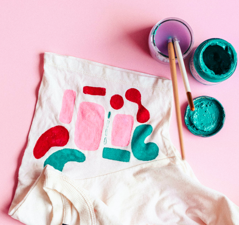 White cloth painted with abstract patterns next to paint and paintbrushes on a pink background
