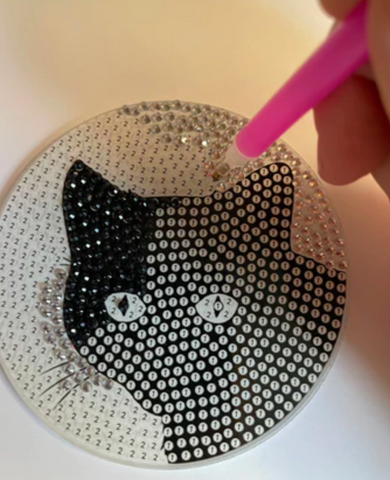 A person adds a jewel to a diamond painted coaster with a cat design