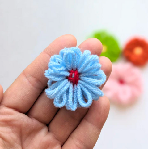 A hand holds a blue and red flower made out of yarn. Three blurred yarn flowers are in the background