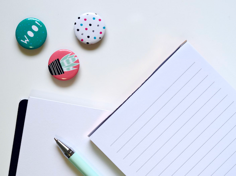 Three button pins with different images: the text "woo!", a polka dot pattern, and a cartoon plant. Next to them are two blank notebooks and a blue pen on a white background.