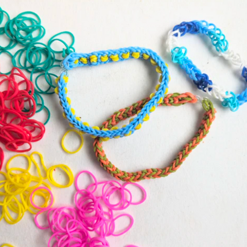 Three rubber band bracelets lay next to piles of small rubber bands in teal, red, yellow, and pink. One bracelet is blue and yellow, another is coral and olive green, and the last is gradients of blue and white. They are on a white background.