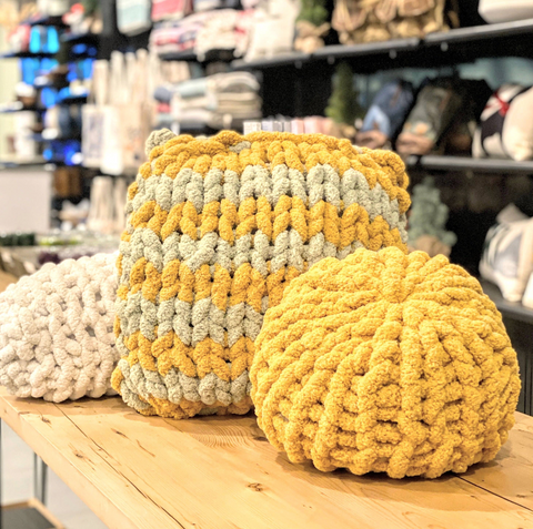 Three chunky knit pillows on a table. One is round and yellow, one square and yellow and green stripes, and one is ivory and an oblong shape.