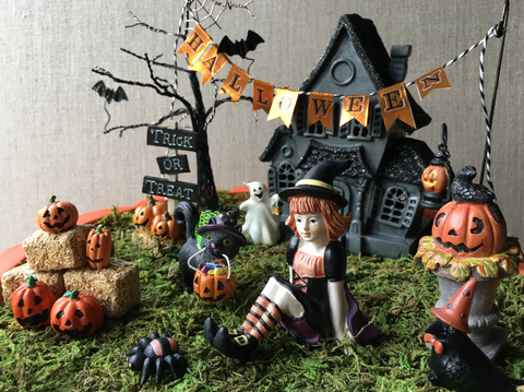 A miniature Halloween scene with pumpkins, a witch, a spooky house, bats, and a tree with no leaves. The figures sit on a bed of moss.